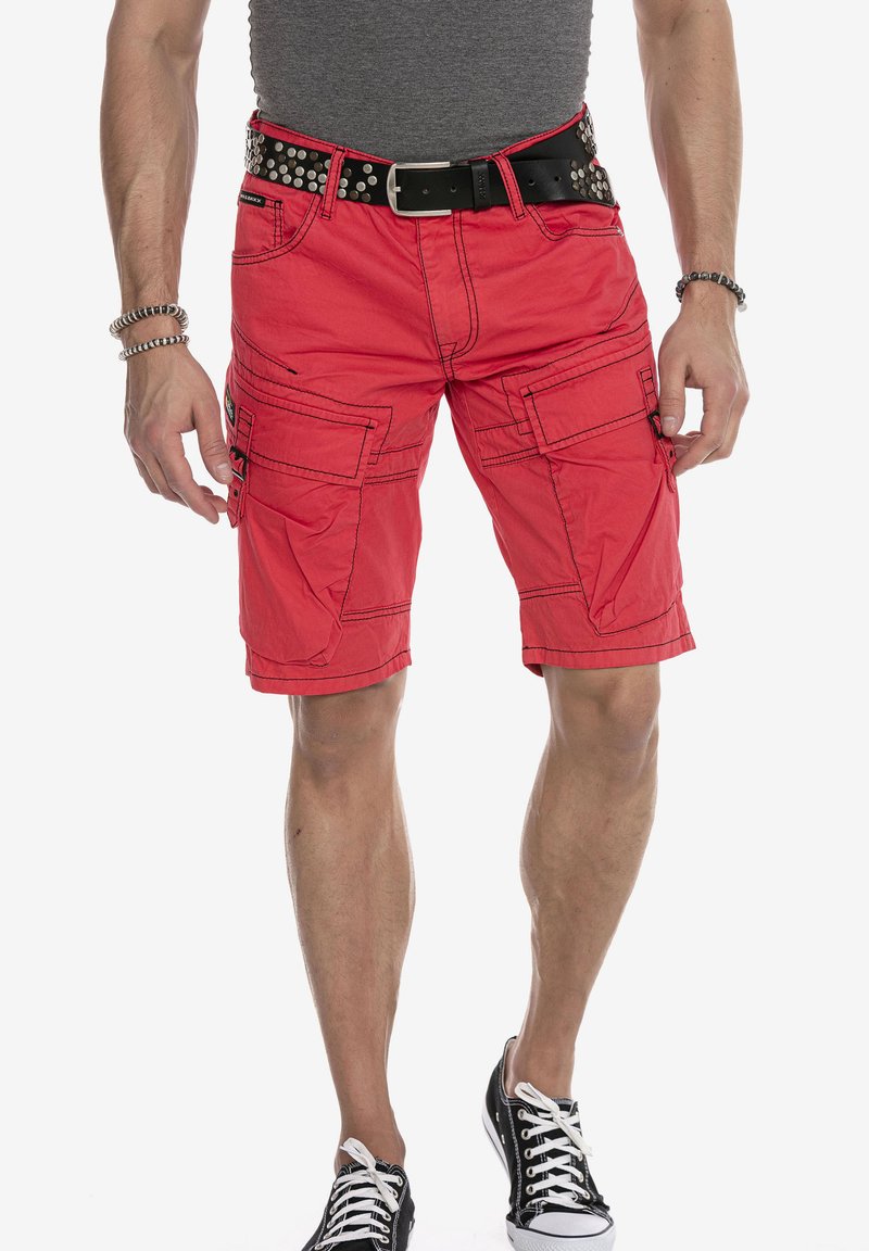 Rote Cargo-Shorts mit mehreren Taschen, Kontrastnähten und einem schwarzen Nieten-Gürtel. Getragen mit einem grauen, enganliegenden Shirt und schwarzen Sneakers.