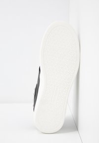 Musta liukujalkine, jossa on tekstuuripintainen päällinen ja pyöreät koristeelliset niitit, sekä valkoinen kumipohja, jossa on geometrinen kuvio.