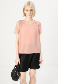 Ροζ μπλούζα σε απαλό χρώμα με κοντά μανίκια από δαντέλα, συνδυασμένη με μια μαύρη φούστα. Η μπλούζα έχει χαλαρή εφαρμογή και στρογγυλή λαιμόκοψη.