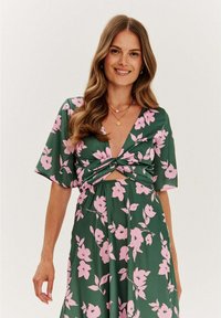 Kvetinové šaty zo zelenej tkaniny s ružovými kvetmi, s krátkymi rukávmi, hlbokým výstrihom do V a uzlovým detailom v páse. Hladká textura.