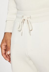 Light beige sweatpants feature a ribbed waistband with a drawstring and side pockets, made from a soft, textured fabric.