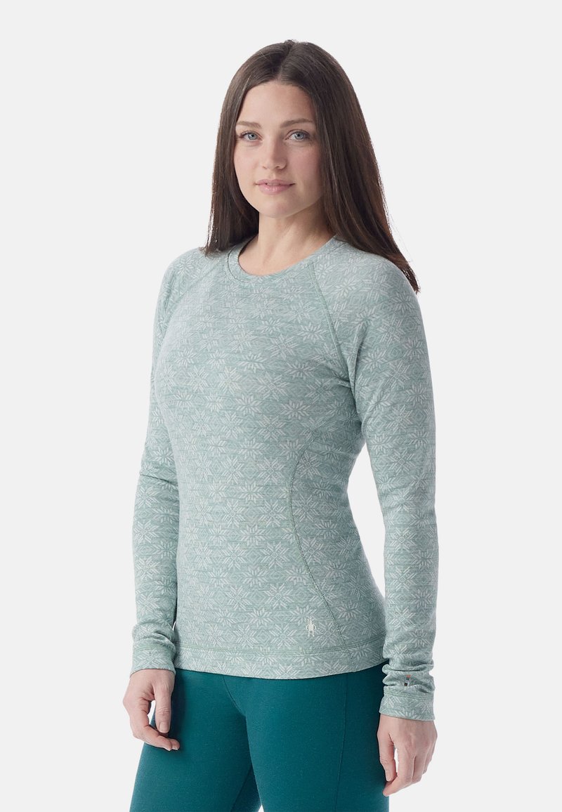 Woman with long brown hair wearing a fitted light green patterned long-sleeve top and teal leggings, standing against a plain background.