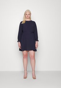 Vestido azul-marinho com mangas longas, decote com botões, cinto na cintura e bainha rodada. Combinado com saltos de tira dourados. Textura suave.