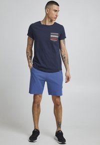 T-shirt navy con tasca a righe, pantaloni corti blu texture e sneakers nere. L'outfit presenta un design moderno e informale, con una vestibilità comoda.