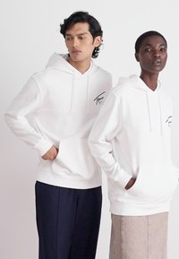 Sudaderas blancas con un bolsillo frontal y cordones presentan un pequeño logo en el pecho. Un modelo lleva pantalones azul marino, el otro en beige.