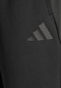 Zwarte sportbroek van gladde stof met een gestructureerd donkergrijs Adidas-logo op de linker dij, met een slanke, taps toelopende pasvorm.