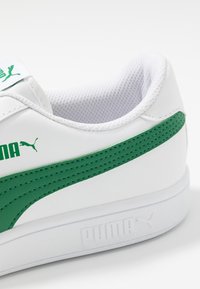 Białe sneakersy z zielonymi akcentami, charakteryzujące się gładką syntetyczną cholewką, teksturowanym panelem bocznym oraz gumową podeszwą z wytłoczonym logo.