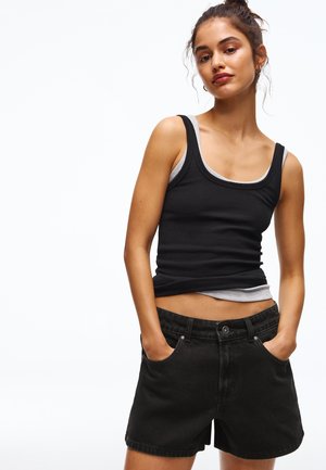 Žena s rukama v kapsách, oblečená v vrstvených černých a šedých tílkách a černých džínových šortkách, stojící před bílým pozadím.