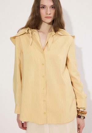 Jeune femme aux longs cheveux bruns portant une chemise ample jaune pâle à boutons avec des détails noués au col et plusieurs bracelets à son poignet.