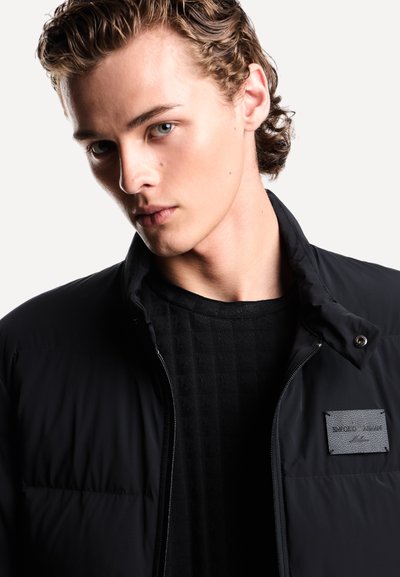 Zwarte pufferjack met een rits, gestructureerde voering en drukknoopkraag. Voorzien van een grijs merkembleem op de borst.