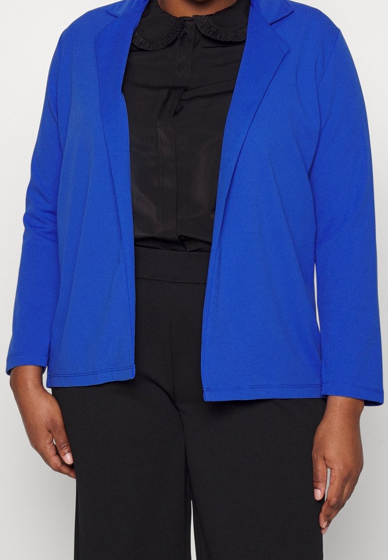 Personne portant un blazer bleu roi ouvert sur un chemisier noir à col volanté, associé à un pantalon noir.