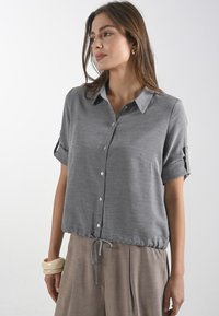 Camicia grigia a maniche corte con colletto a bottoni, dotata di orlo con cordoncino e una tasca singola, realizzata in un tessuto morbido.