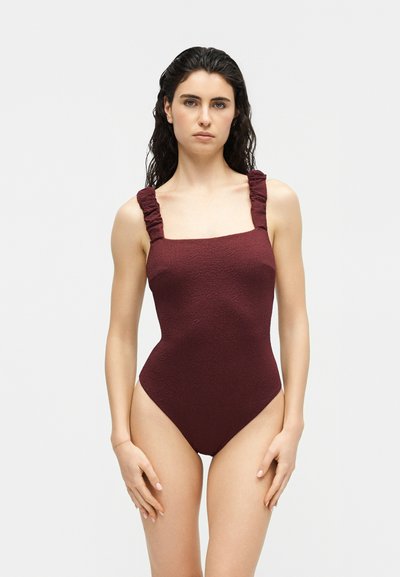 Femme aux cheveux bouclés foncés portant un maillot de bain une pièce bordeaux texturé avec des bretelles froncées, debout devant un fond uni.