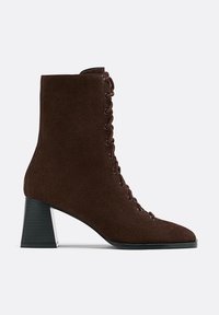 Stivale alla caviglia in suede marrone con punta affusolata, design all'altezza della caviglia, dettagli di allacciatura evidenti e un tacco a blocco spesso accentuato da una rifinitura argentata.
