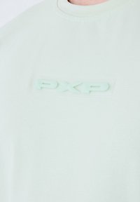 T-shirt di cotone verde chiaro con colletto a girocollo e logo "PXP" in rilievo in un materiale traslucido sulla parte anteriore. Tessuto liscio, design casual.