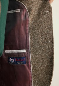 Brown tweed jacket interior features a burgundy herringbone lining, white pocket trim, and a label reading "Shetland, Woven in England, Since 1837."
