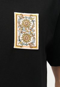 Κεντημένο έμβλημα σε μαύρο ύφασμα που απεικονίζει δύο περίτεχνους χρυσούς ήλιους με πρόσωπα, περιτριγυρισμένους από μπλε και πορτοκαλί διακοσμητικά φυλλώματα.