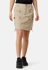 Beige functional skirt with two front zippered pockets and side zippers, crafted from lightweight material. Worn with black athletic shoes.