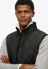 Chaleco negro acolchado con cuello alto, que presenta botones negros y un patrón texturizado. Llevado sobre una camiseta de manga larga gris.