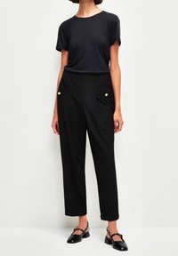 Musta t-paita käännetyillä hihoilla, jotka on yhdistetty mustiin korkeavyötäröisiin housuihin, joissa on kultaiset nappikoristeet ja sivutaskut. Mustat tasapohjaiset kengät täydentävät asua.