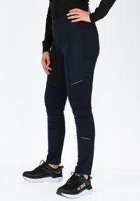 Leggings atletici blu marine con una finitura testurizzata, caratterizzati da tasche laterali con zip e dettagli dorati, abbinati a scarpe sportive nere.
