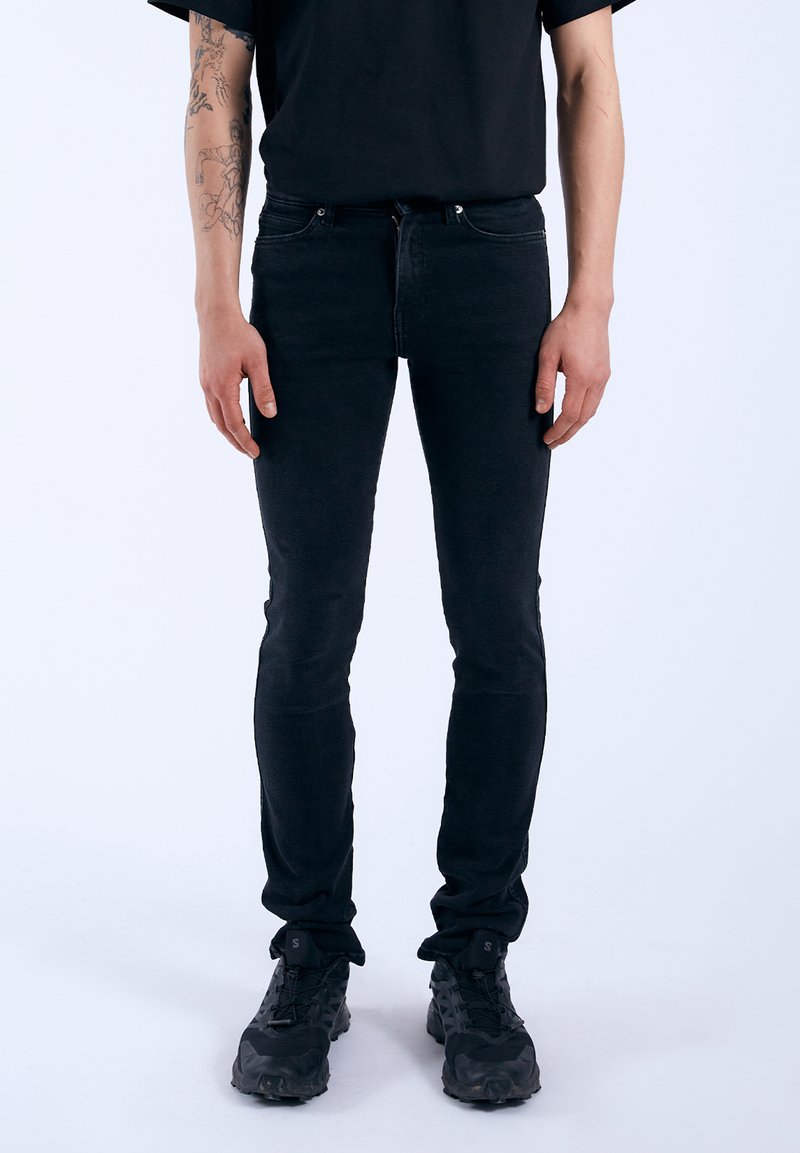 Vaqueros negros ajustados de denim, con un diseño clásico, cinco bolsillos y cremallera. Combinados con calzado negro texturizado.