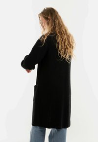 Vrouw met lang krullend haar, gekleed in een zwarte gebreide cardigan met zakken en blauwe jeans, staande in profiel tegen een witte achtergrond.