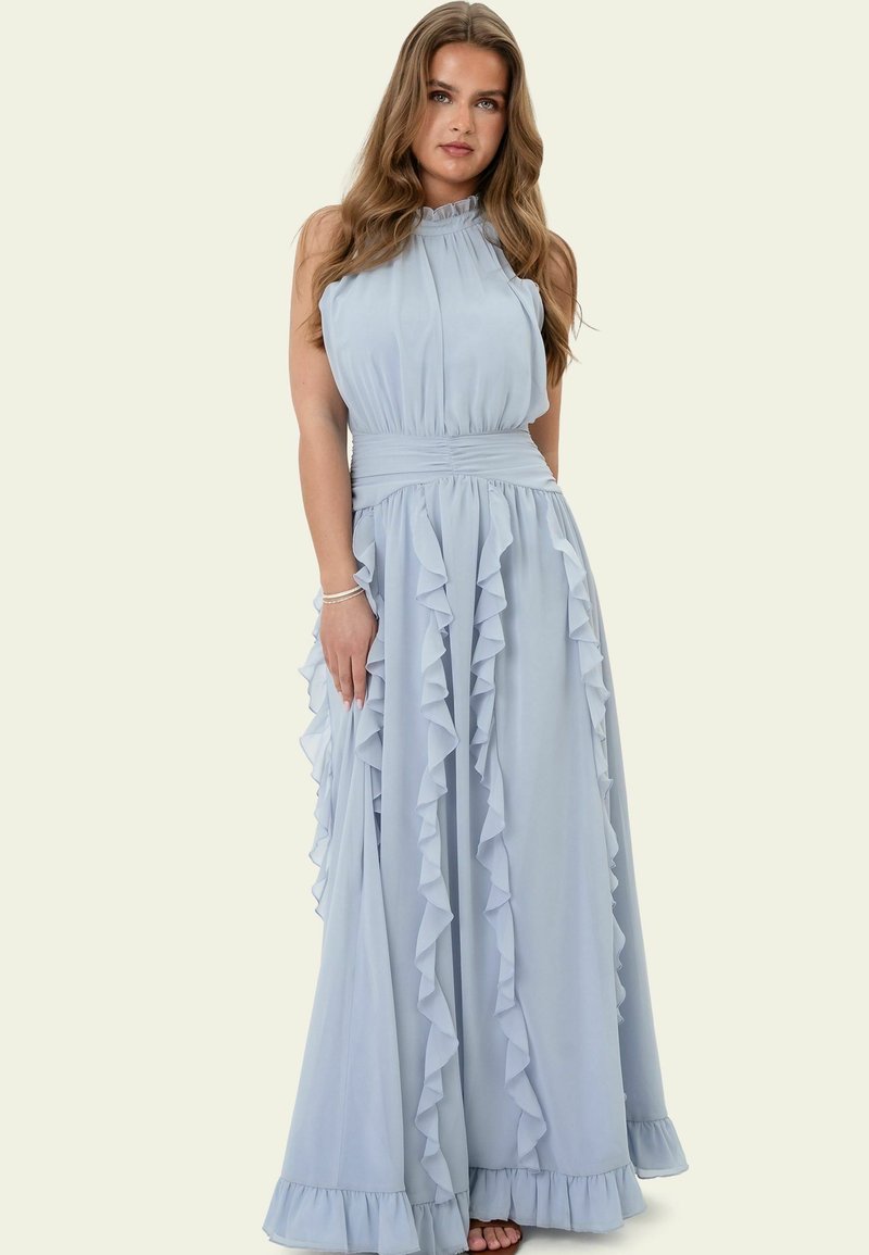 Femme aux longs cheveux ondulés portant une robe maxi bleu clair sans manches avec des volants et un col haut, debout devant un fond uni.