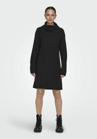 Robe noire en tricot avec un col drapé, un motif texturé, des manches longues et une coupe droite, associée à des bottes à lacets noires à semelle épaisse.