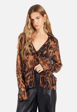 Młoda kobieta z długimi blond włosami w przezroczystej brązowo-czarnej bluzce w panterkę i czarnych skórzanych spodniach, pozująca na białym tle.