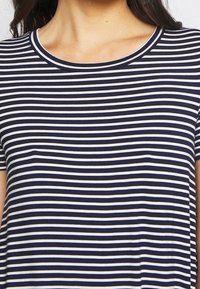 Kobieta ubrana w granatową i białą, poziomo paskowaną koszulę z krótkim rękawem i okrągłym dekoltem, pokazana od szyi do środka torsu.