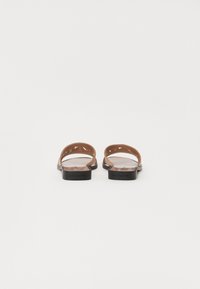 Sandales en cuir marron avec deux sangles courbées, arrière ouvert, semelles plates noires, présentant des motifs triangulaires découpés sur les sangles.