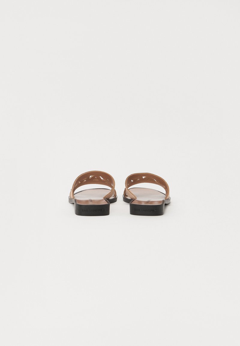Sandales en cuir marron avec deux sangles courbées, arrière ouvert, semelles plates noires, présentant des motifs triangulaires découpés sur les sangles.