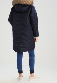 Námořnická modrá parka s kapucí lemovanou kožešinou, s uzávěrem na zip a druky, bočními kapsami a mírně staženým pasem pro lepší tvar.