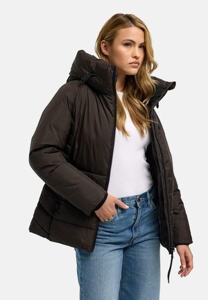 Woman with long blonde hair wearing a black puffer jacket, white shirt, and blue jeans, standing with one hand in pocket.