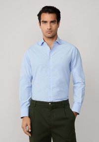Camisa de botones de manga larga color azul claro, hecha de tela de algodón, con un cuello clásico y detalles bordados, combinada con pantalones de color verde oscuro.
