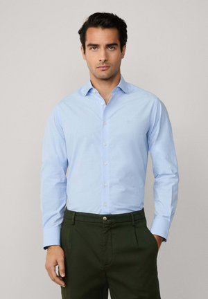 Camisa de botones de manga larga color azul claro, hecha de tela de algodón, con un cuello clásico y detalles bordados, combinada con pantalones de color verde oscuro.