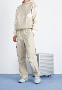 Felpa beige con disegno bianco; pantaloni cargo larghi e leggeri in una tonalità di beige più chiara; scarpe sportive grigie con dettagli scuri.