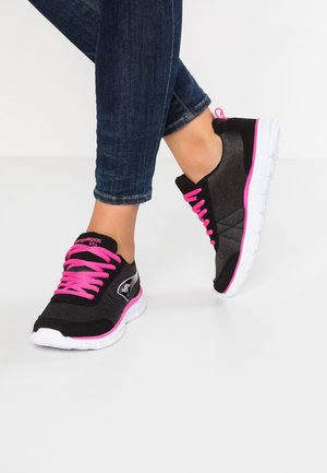 Czarne i różowe buty sportowe z białymi podeszwami noszone z ciemnoniebieskimi dżinsami, pokazane na skrzyżowanych nogach na białym tle.