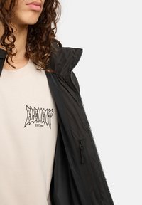 Persona con capelli ricci che indossa una t-shirt grafica beige DLEMNNT e una giacca nera parzialmente aperta, inquadrata dalle spalle al petto.