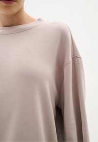 Femme portant un haut taupe à manches longues amples avec épaules tombantes et coutures visibles sur les manches et l'encolure.