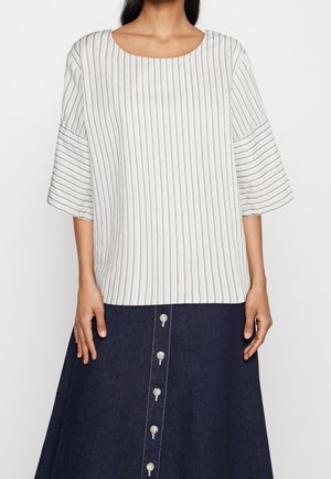 Vrouw draagt een witte blouse met dunne verticale strepen en wijde mouwen, gecombineerd met een donkere rok met knopen.