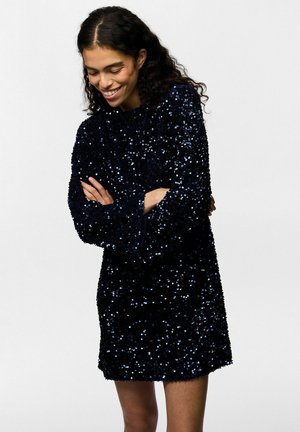 Mujer con cabello oscuro y rizado que lleva un vestido negro de manga larga con lentejuelas, de pie con los brazos cruzados y sonriendo mientras mira hacia abajo.