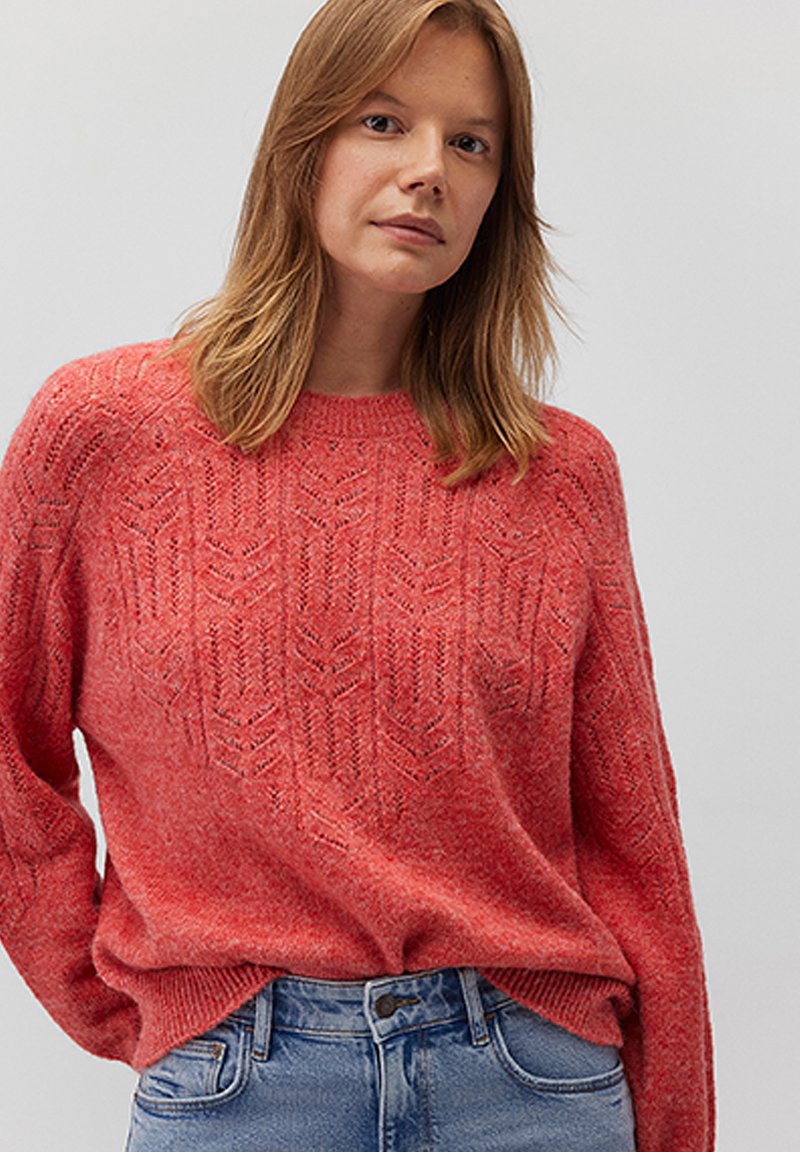 Roter gestrickter Pullover mit strukturiertem Muster, rundem Ausschnitt und gerippten Bündchen. Kombiniert mit hellblauen Jeans.