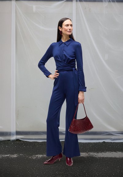 Marineblå jumpsuit med omslagsdesign, lange ærmer og knapdetaljer; kombineret med en burgunderflettet håndtaske og burgunder hæle.
