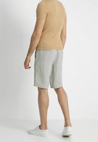 Beżowa, prążkowana koszula z krótkim rękawem w połączeniu z jasnoszarymi, lnianymi szortami. Model ma na sobie białe tenisówki. Widok z boku, minimalistyczne tło.