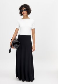 Zwarte maxi rok en witte top met korte mouwen. De rok heeft een vloeiende uitstraling; het model houdt een zwarte clutch vast en draagt donkere zonnebrillen.