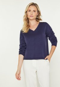 Pulover bleumarin cu decolteu în V, mâneci lungi, tricot cu textură, cu mansete și tiv ribate, asortat cu pantaloni albi. Croială simplă, modernă.