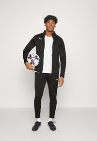 Mustat urheilu takki ja housut, joissa valkoiset Puma-logot, valkoinen t-paita ja mustat nappulakengät. Pitelee moniväristä jalkapalloa. Minimalistinen tausta.