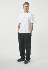 Weißes T-Shirt mit kurzen Ärmeln aus Baumwolle, kombiniert mit schwarzen, lässig sitzenden Hosen. Benutzer trägt schwarze Sportschuhe und steht mit den Händen in den Taschen.